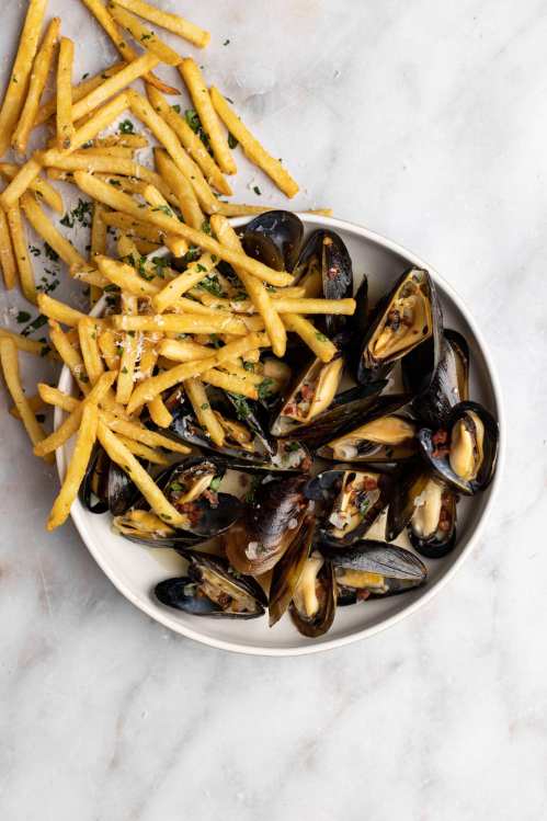 A bowl of mussels with garlic and herbs, served alongside crispy French fries on a marble surface.