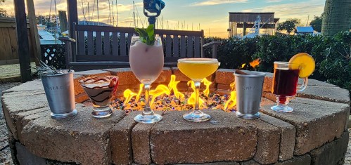 Five colorful cocktails arranged around a fire pit, with a sunset and boats in the background.