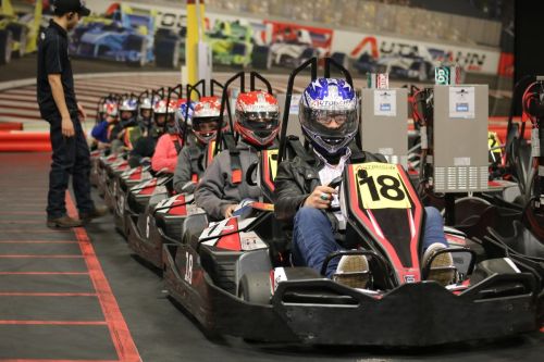 A line of go-karts with drivers wearing helmets, ready to race on an indoor track.