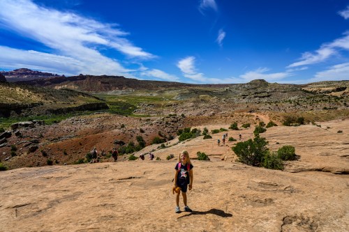 A child stands on rocky terrain with a vast landscape and blue sky in the background, surrounded by other hikers.
