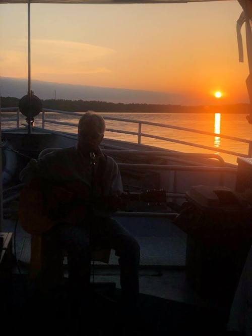 A musician plays guitar on a boat at sunset, with a serene water view and warm colors in the sky.