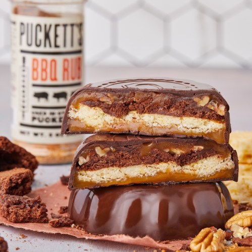 A close-up of stacked chocolate bars with layers of caramel, cookie, and brownie, surrounded by nuts and BBQ rub.