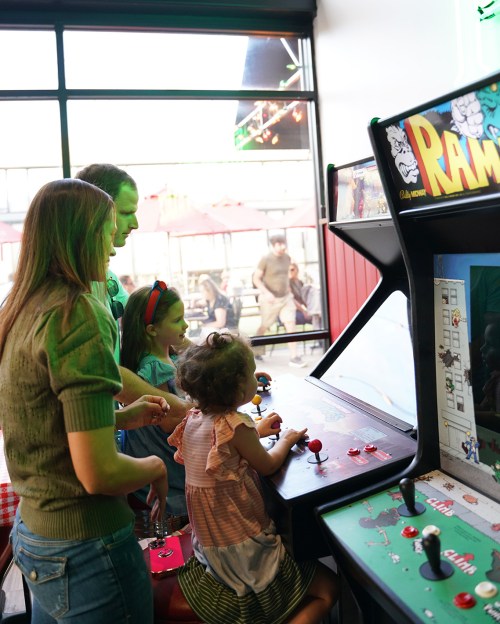 A family enjoys playing an arcade game together, with two children focused on the controls.