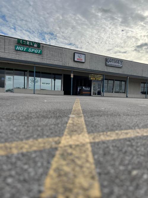 A storefront with signs for "Kelly's Hot Spot" and "Odyssey," under a cloudy sky and a parking lot view.
