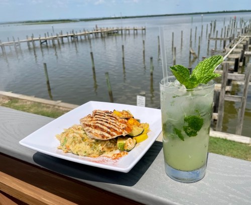 A plate of grilled fish and vegetables beside a refreshing mint mojito, with a scenic waterfront view in the background.