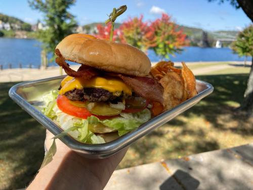 A hand holding a cheeseburger with bacon, lettuce, tomato, and pickles, alongside crispy potato chips, by a scenic river.