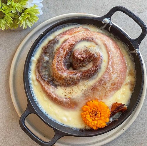A cinnamon roll in a cast iron skillet, topped with creamy icing and a marigold flower, on a light-colored plate.