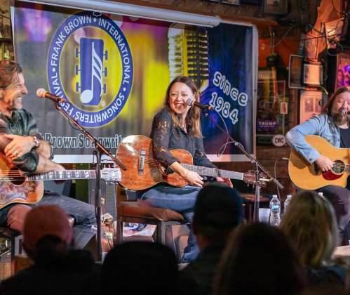 A live music performance featuring three musicians with guitars, smiling and engaging with the audience in a cozy venue.