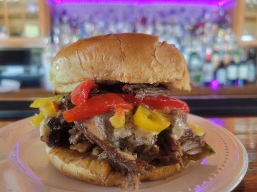A hearty sandwich with pulled beef, peppers, and sauce on a bun, served on a plate with a blurred bar background.