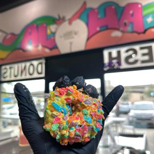 A colorful donut topped with fruity cereal, held in a gloved hand, with a vibrant mural in the background.