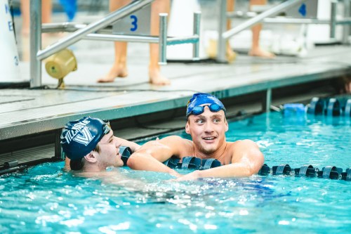 Two male swimmers in a pool, smiling and chatting while resting against the pool edge.