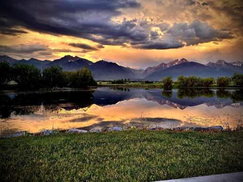 A serene landscape featuring mountains reflecting in a calm lake at sunset, with vibrant clouds and greenery in the foreground.