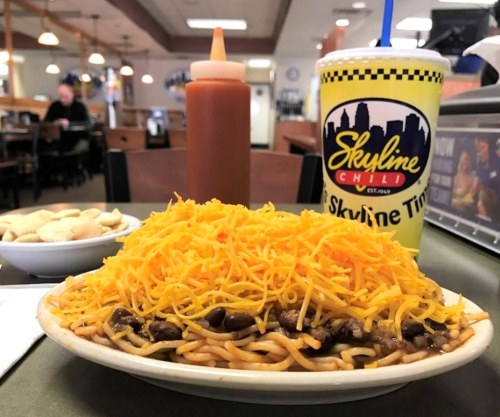 A plate of spaghetti topped with chili and a mound of shredded cheese, with a Skyline drink and condiments in the background.
