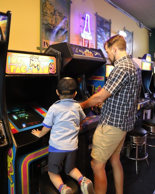 A man and a child play arcade games together, focused on the screens in a colorful game room.