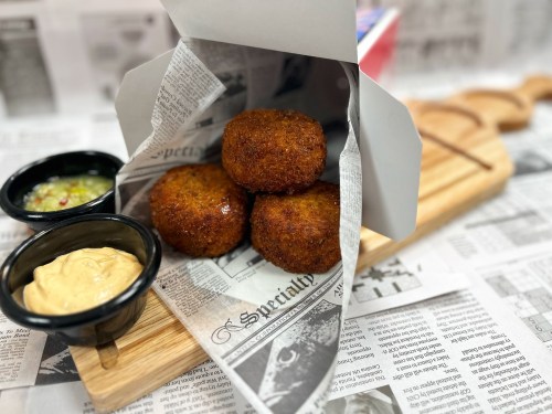 Three golden-brown croquettes in a paper cone, served with two dipping sauces on a newspaper backdrop.