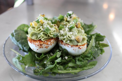 Two avocado-filled deviled eggs topped with greens and cheese, served on a bed of fresh spinach.