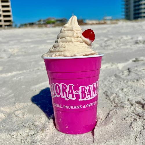 A pink cup filled with soft serve ice cream topped with a cherry, sitting on sandy beach with buildings in the background.