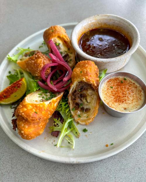 A plate with crispy egg rolls, dipping sauces, pickled onions, and a lime wedge, garnished with greens.