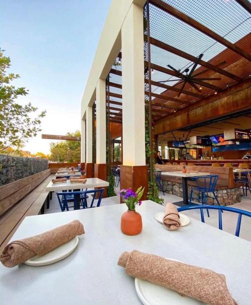 A modern restaurant patio with tables set for dining, featuring greenery and a stylish interior in the background.