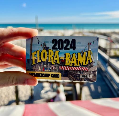 A hand holds a 2024 Flora-Bama membership card against a beach backdrop with blue skies.