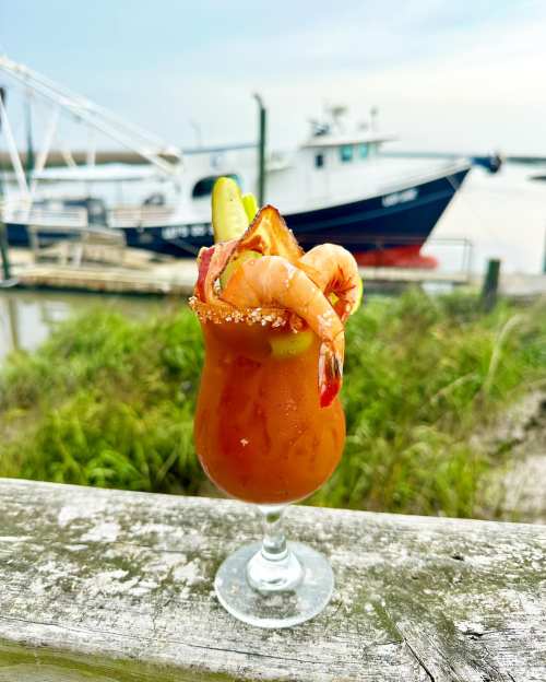 A vibrant cocktail garnished with shrimp, cucumber, and pineapple, set against a scenic waterfront backdrop.