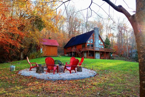 A cozy red cabin surrounded by autumn foliage, featuring a fire pit with red chairs in a scenic outdoor setting.