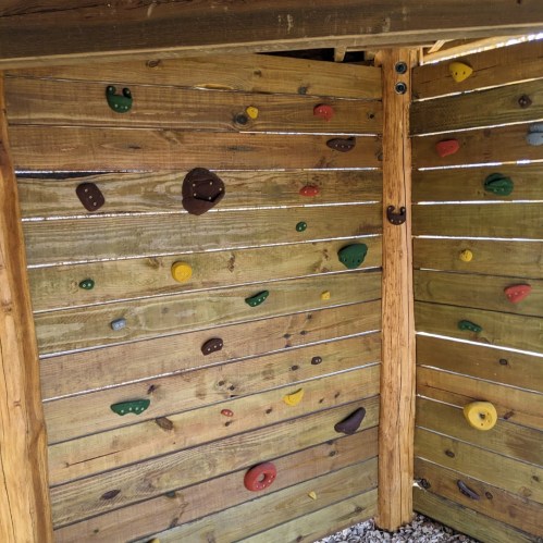 A wooden climbing wall with colorful holds in various shapes, set in a rustic outdoor environment.