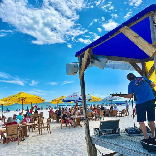 A musician performs on a beach stage with umbrellas and seated guests enjoying the sunny day.