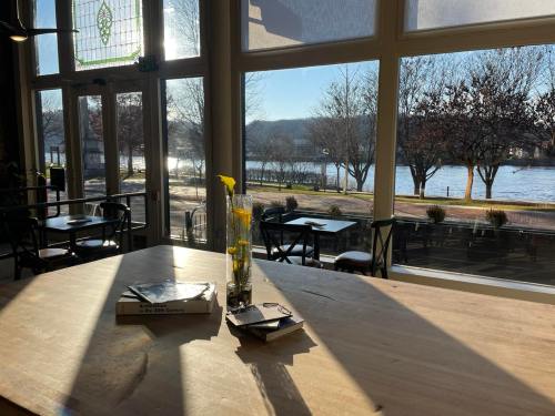 A sunlit interior with a wooden table, books, and a vase, overlooking a river and trees through large windows.