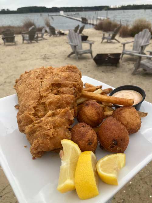 A plate of fried fish, fries, and hushpuppies with lemon wedges, set against a beach and water backdrop.