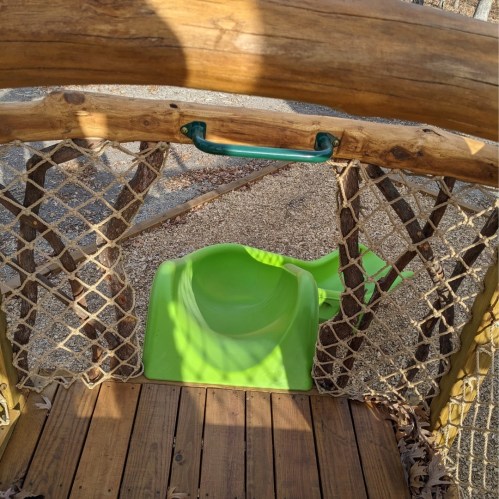 A wooden playground structure with a green slide and a rope netting, set on a sandy surface.