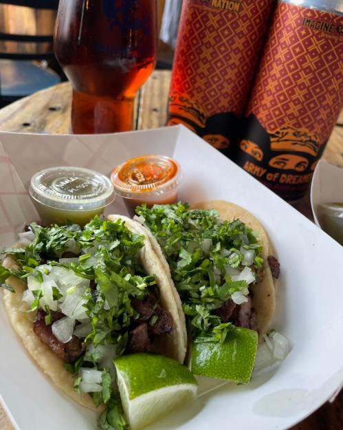 Two tacos filled with meat, topped with onions and cilantro, served with lime wedges and sauces, alongside a drink.