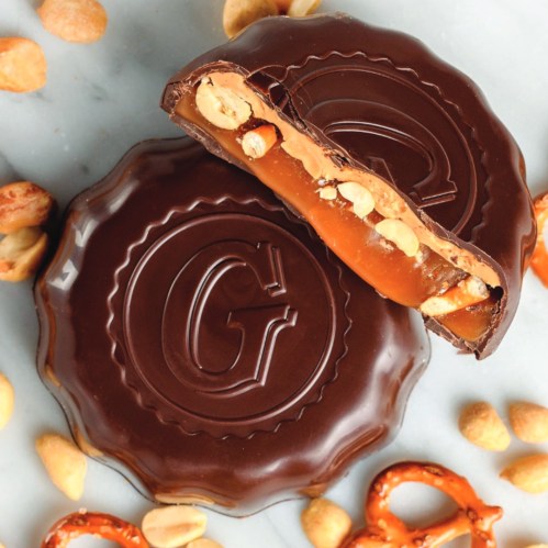 A chocolate-covered treat with a caramel and peanut filling, surrounded by peanuts and pretzels.