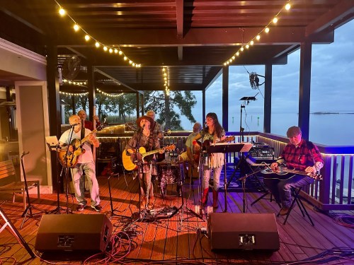 A live band performs on a deck with string lights, overlooking a body of water, creating a vibrant atmosphere.