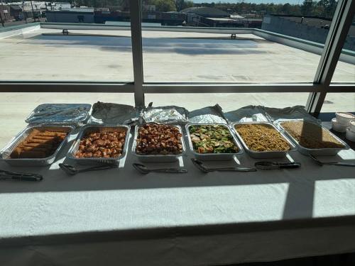 A table with various food trays, including chicken, vegetables, noodles, and spring rolls, set against a window view.