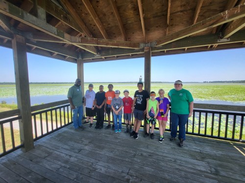 A group of ten people stands on a wooden deck overlooking a scenic wetland with water and lily pads.
