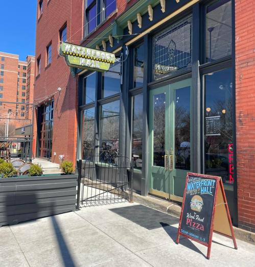 Exterior of Waterfront Hall with a sign for wood-fired pizza and a welcoming entrance on a sunny day.