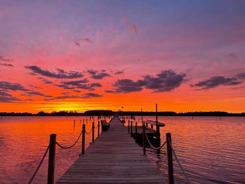 A wooden dock extends into a calm lake at sunset, with vibrant orange and purple skies reflecting on the water.