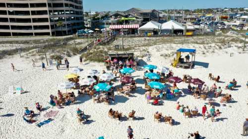 A beach scene with people sitting under umbrellas, a boardwalk, and nearby buildings in a sunny atmosphere.