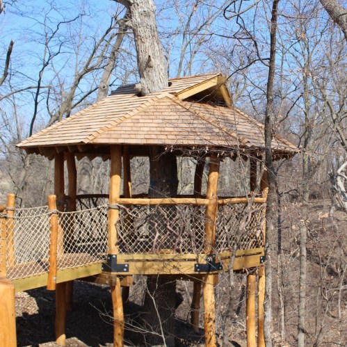 A wooden treehouse with a shingled roof, surrounded by bare trees and a wooden railing.