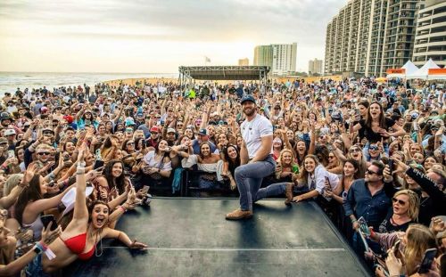 A lively beach concert scene with a large crowd cheering and a performer on stage, set against a sunset backdrop.