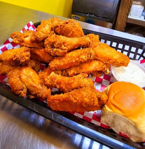 A basket of crispy fried chicken wings with a side of a soft bun and dipping sauce on a checkered paper.