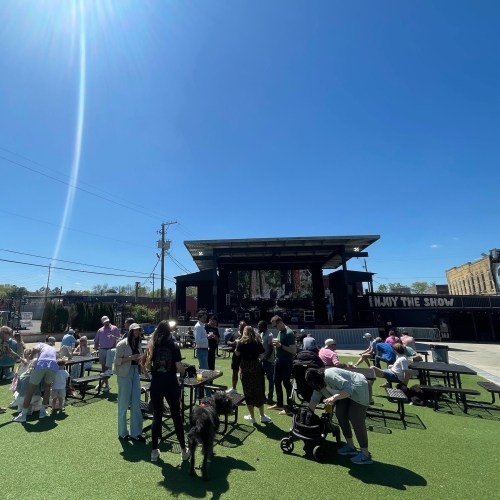 A lively outdoor event with people gathered around tables, a stage in the background, and a clear blue sky above.