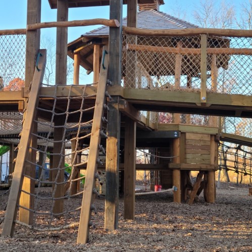 A wooden playground structure with a climbing net and a lookout tower, surrounded by trees and gravel ground.