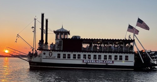 A riverboat named River Lady sails on water at sunset, with American flags waving and people on the upper deck.