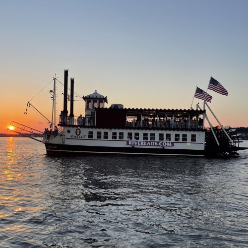 A riverboat named River Lady sails at sunset, with flags waving and passengers enjoying the view.