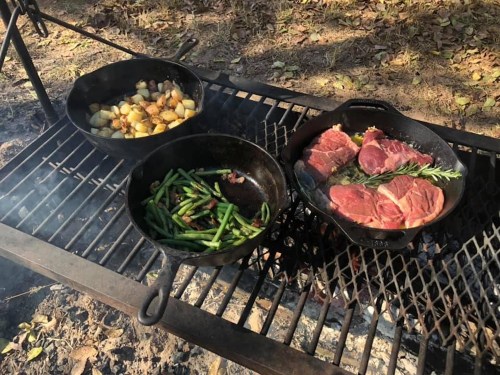 Three cast iron pans on a grill over an open fire, cooking potatoes, green beans, and steaks with rosemary.
