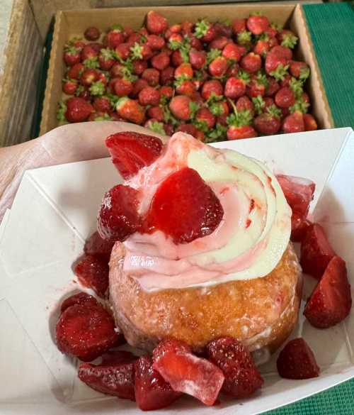 A hand holds a dessert topped with strawberries and cream, with a box of fresh strawberries in the background.