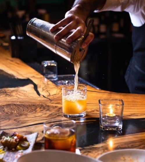 A bartender pours a cocktail from a shaker into a glass on a wooden bar, with candles and food in the background.
