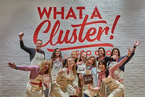 A group of cheerful people in aprons pose in front of a colorful "What a Cluster!" mural, celebrating together.